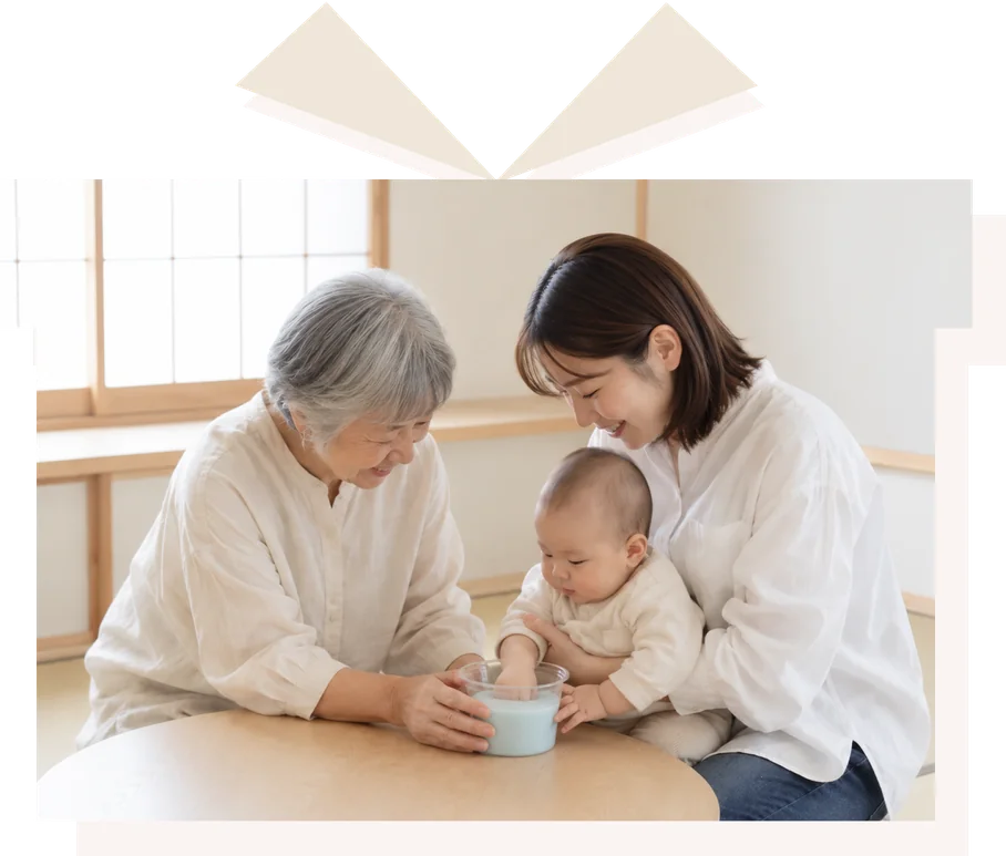 畳の部屋で、祖母と母が赤ちゃんの手を、型取り材の入った透明なカップにそっと入れている様子。リボン付きのギフトボックスの形に切り抜いた写真