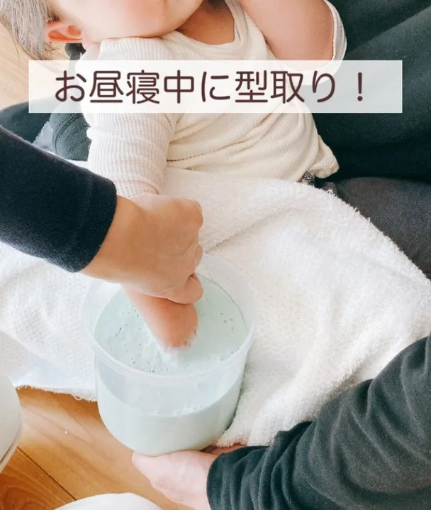 眠っている赤ちゃんの手を、そっと型取り材のカップに入れている様子。お昼寝中に型取り！というリール投稿画像