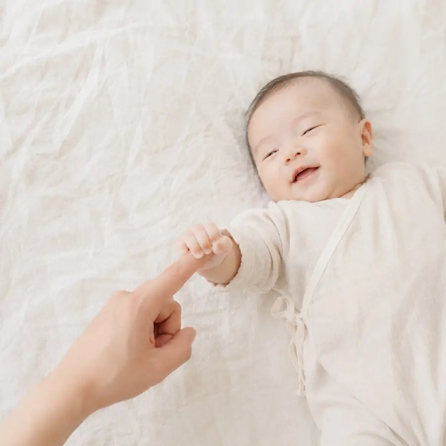 白いリネンの上で、赤ちゃんが大人の人さし指をぎゅっと握って笑っている様子